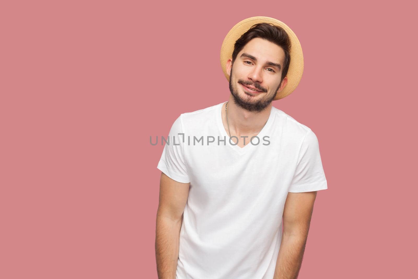 Portrait of happy bearded young man in white shirt with hat standing and looking at camera with smile. indoor studio shot, isolated on pink background copyspace.