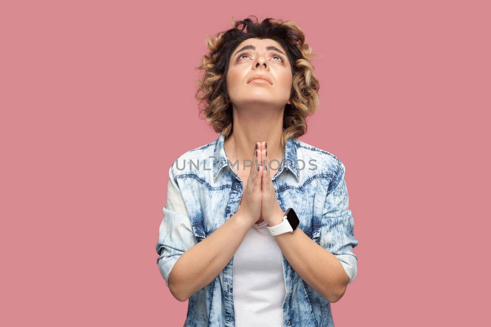 My god please help me. Portrait of hopeful young woman with curly hairstyle in casual blue shirt standing with palm hand and praying or begging. indoor studio shot, isolated on pink background.