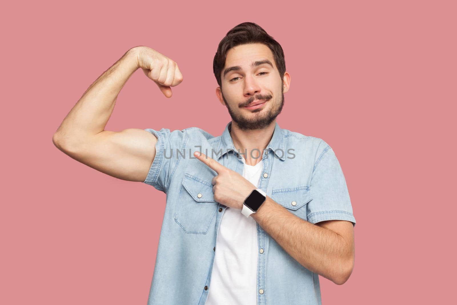 I am strong and I can do anything. Portrait of proud handsome bearded young man in blue casual shirt standing pointing and showing his strong bicep. indoor studio shot, isolated on pink background.