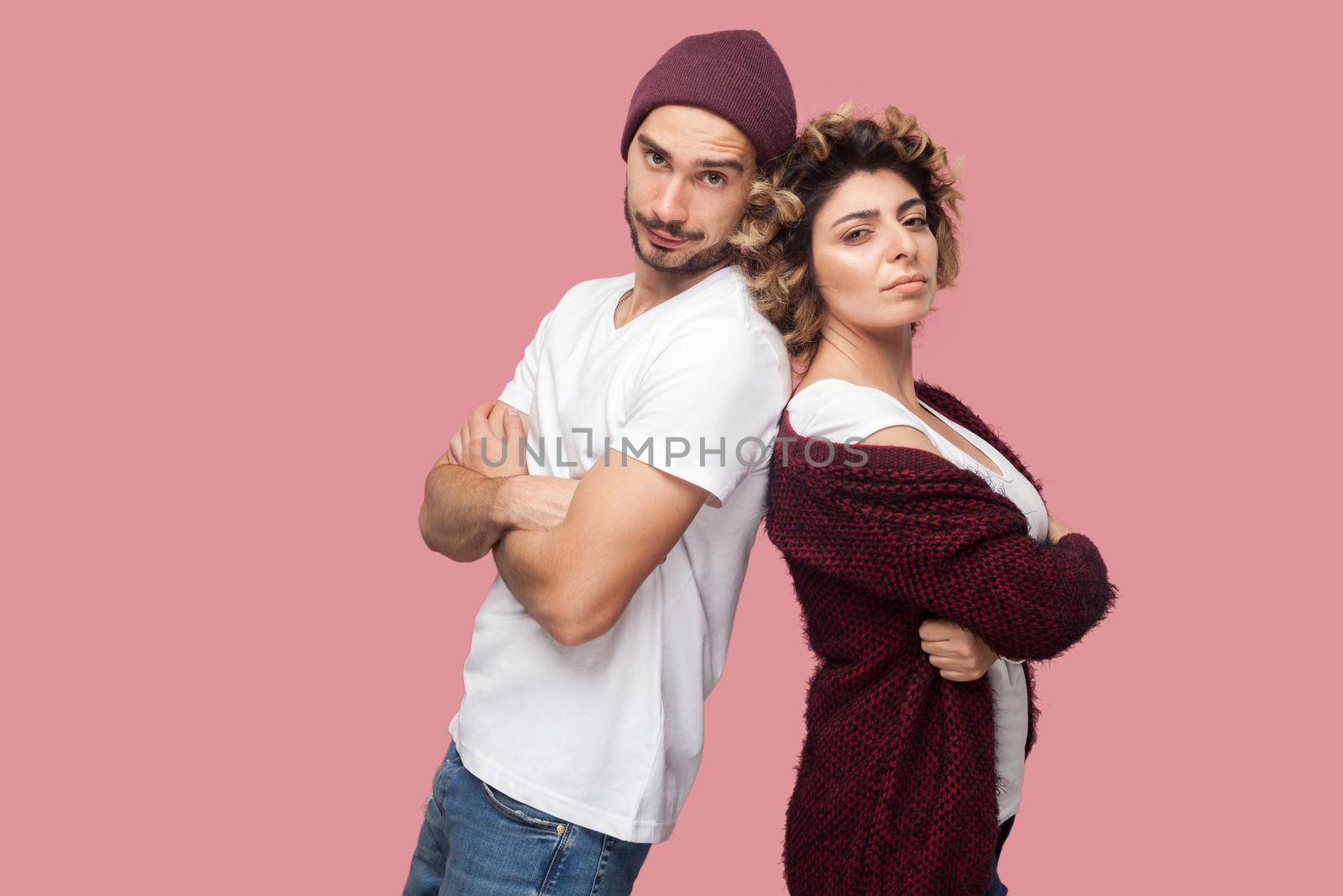 Portrait of serious couple of friends in casual style standing back to back, crossing arms and looking at camera with proud face. Isolated, indoor, studio shot, pink background