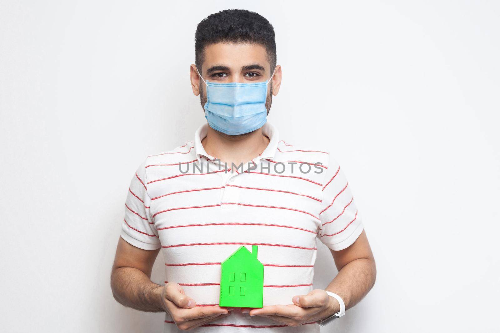 Stay at home, stay safe. Positive man with surgical medical mask holding green small house, looking at camera kindly, reassuring face on quarantine time. Indoor isolated, studio shot, white background