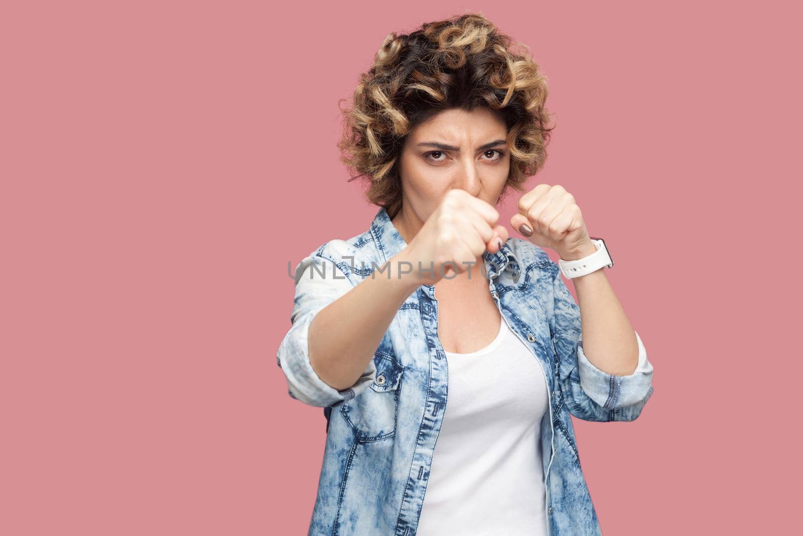 Portrait of serious young woman with curly hairstyle in casual blue shirt standing with boxing fists, looking at camera and ready to attack. indoor studio shot, isolated on pink background.