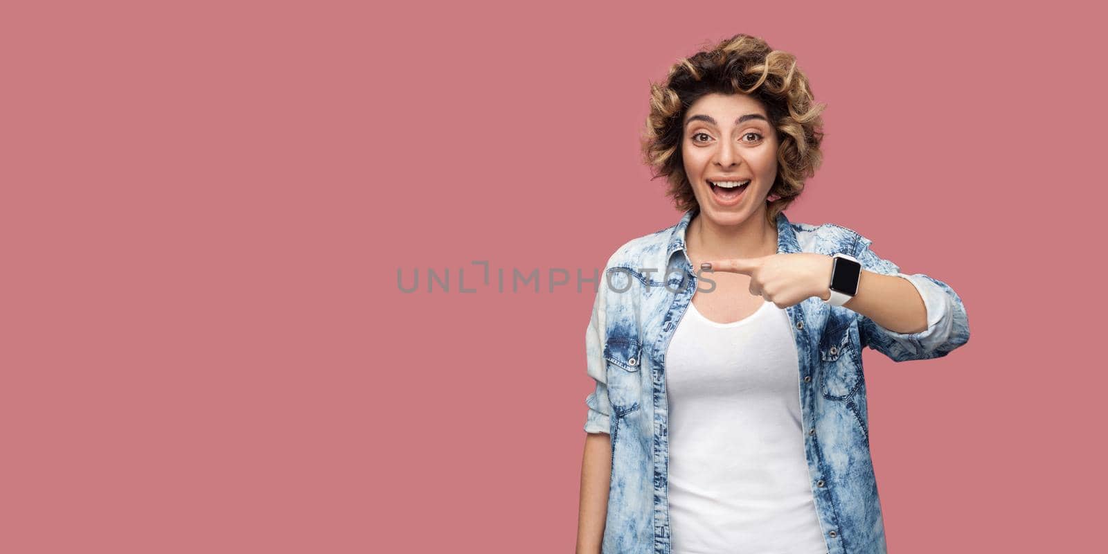 Portrait of amazed young woman with curly hairstyle in casual blue shirt standing and pointing at background empty copyspace with surprised face. indoor studio shot, isolated on pink background.