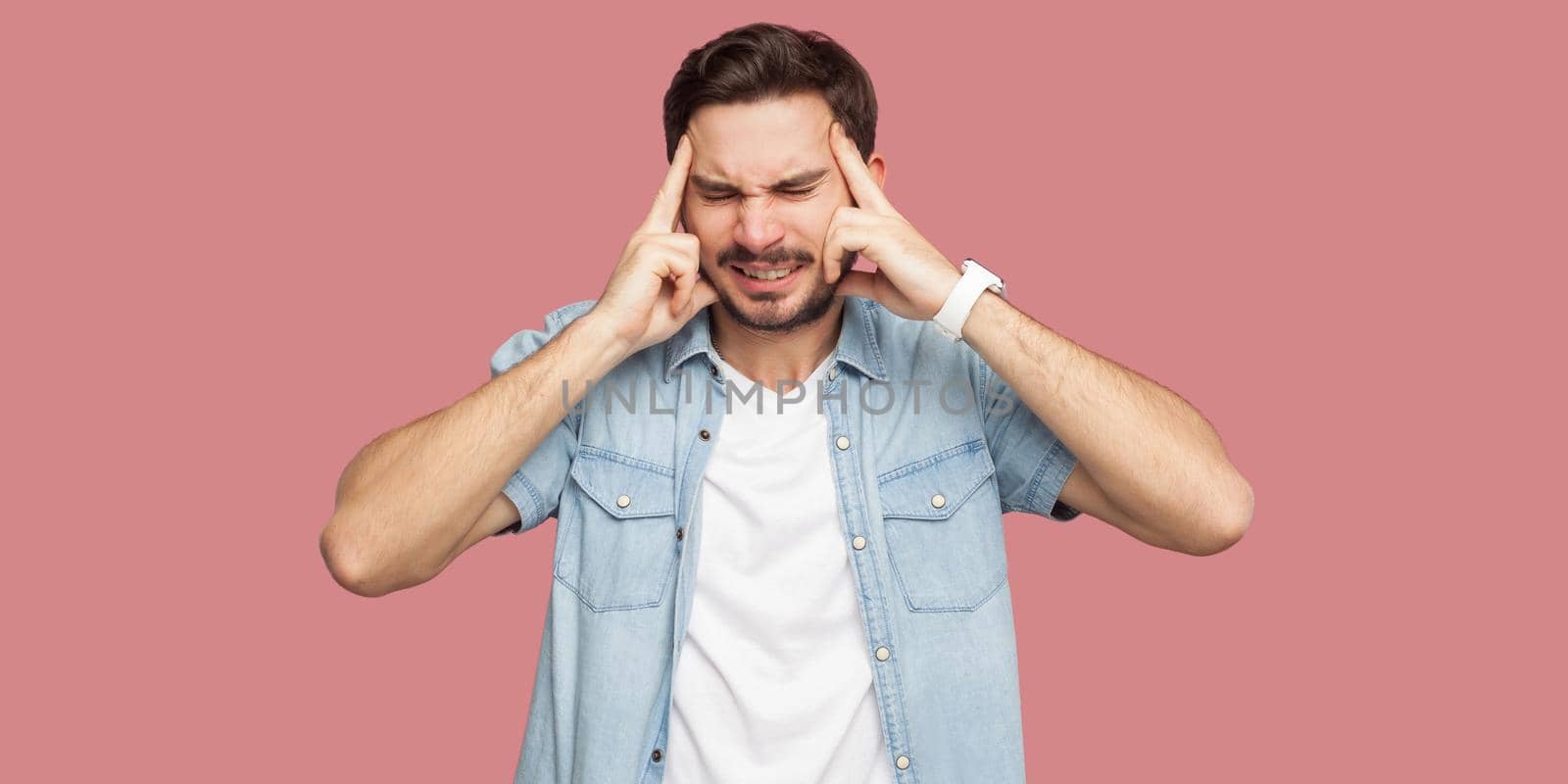 Headache, confusion or thinking. Portrait of sad handsome bearded young man in blue casual shirt standing and holding his head, thinking or endure. indoor studio shot, isolated on pink background.