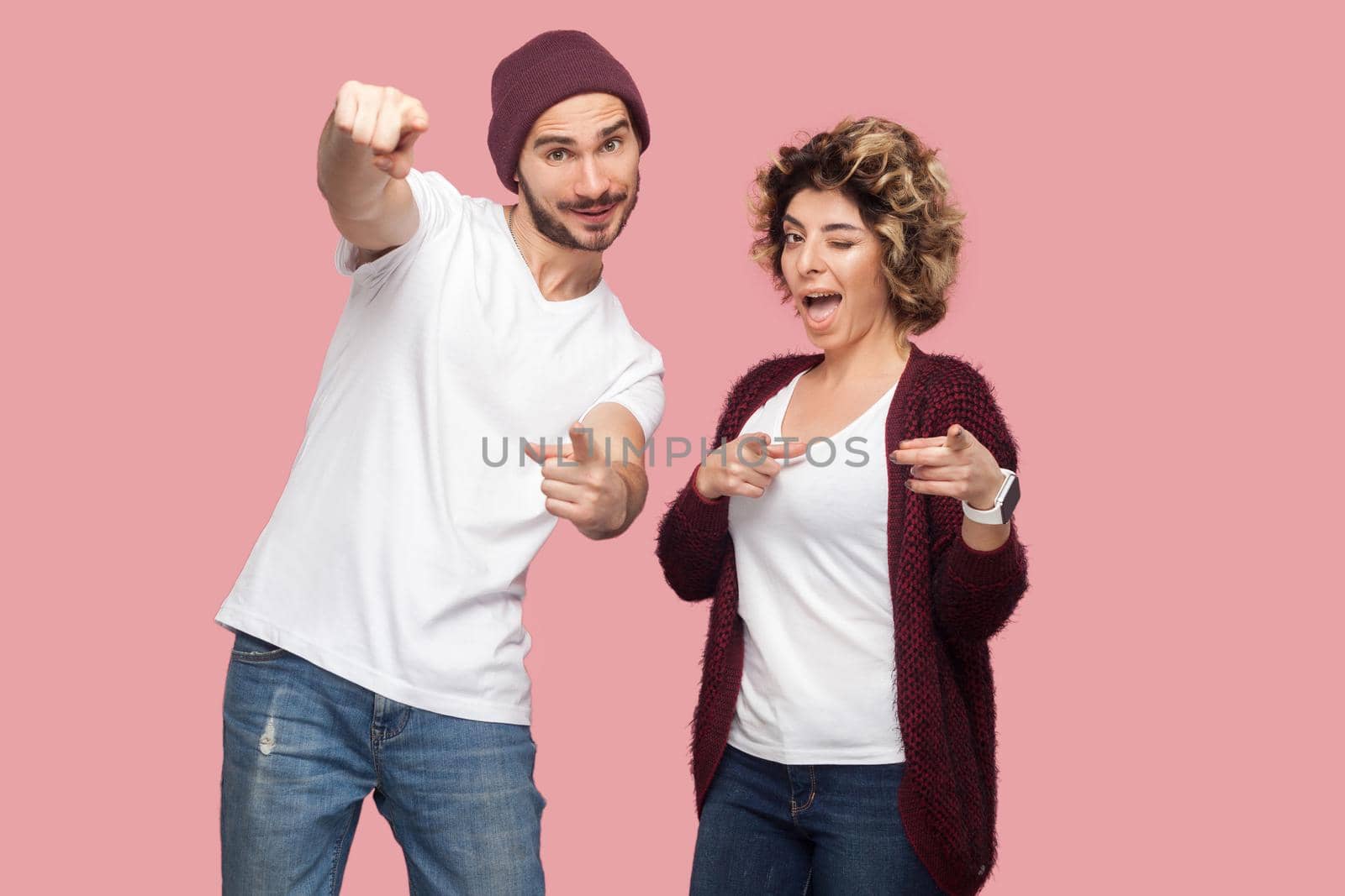 Portrait of funny suprised couple of friends in casual style standing and pointing figrer to you, looking at camera and winking. Isolated, indoor, studio shot, pink background