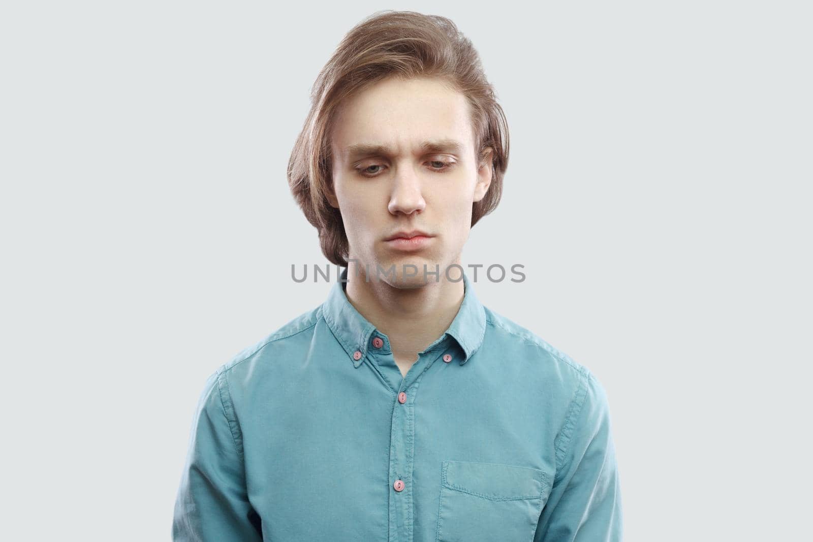 closeup portrait of sad worried handsome long haired blonde young man in blue casual shirt standing, holding head down and thinking what to do. indoor studio shot, isolated on light grey background.