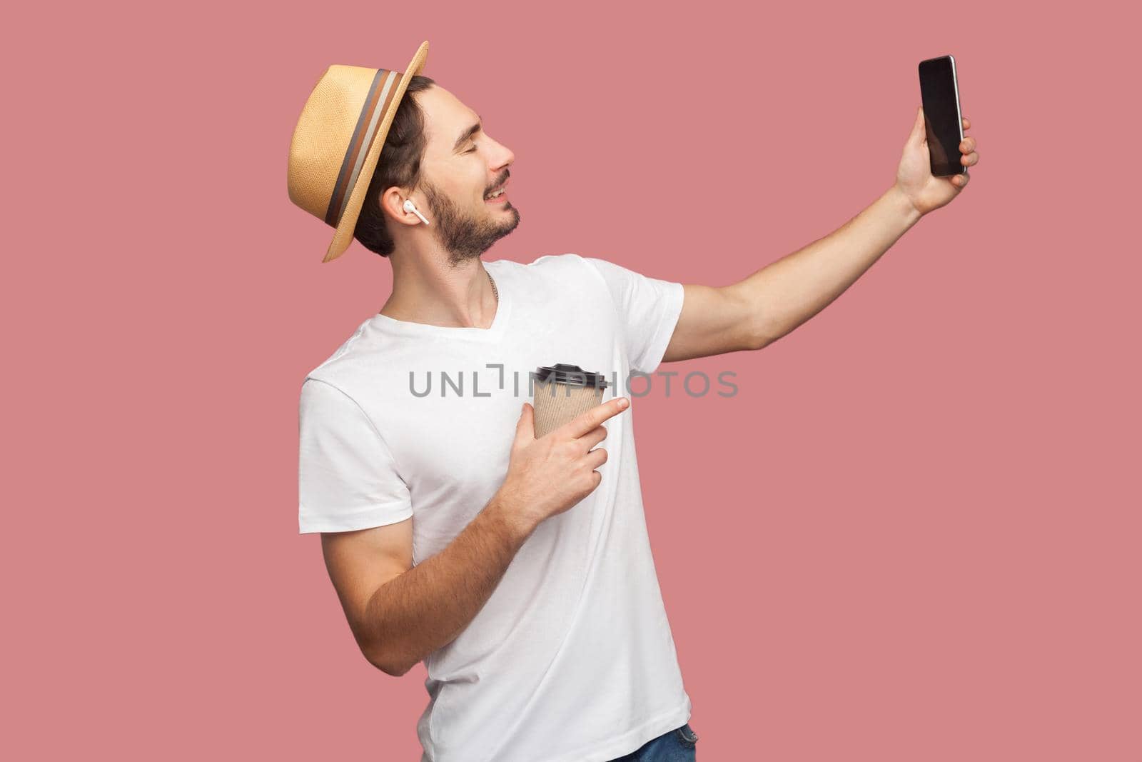 Side view portrait of handsome bearded young hipster man in white shirt and casual hat posing and making selfie photo with smile, blogger lifestyle. Indoor,isolated, studio shot, pink background