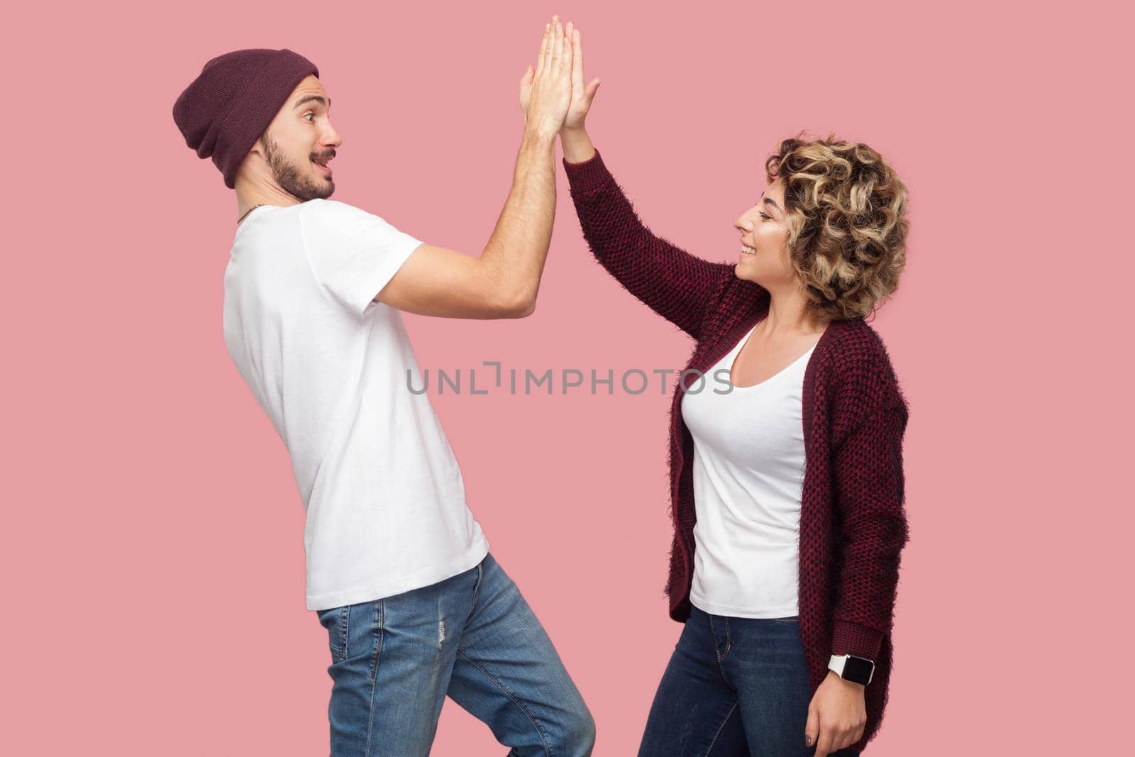Side view portrait of happy couple of friends in casual style standing and giving five to each other with toothy smile, celebrating victory. Isolated, indoor, studio shot, pink background