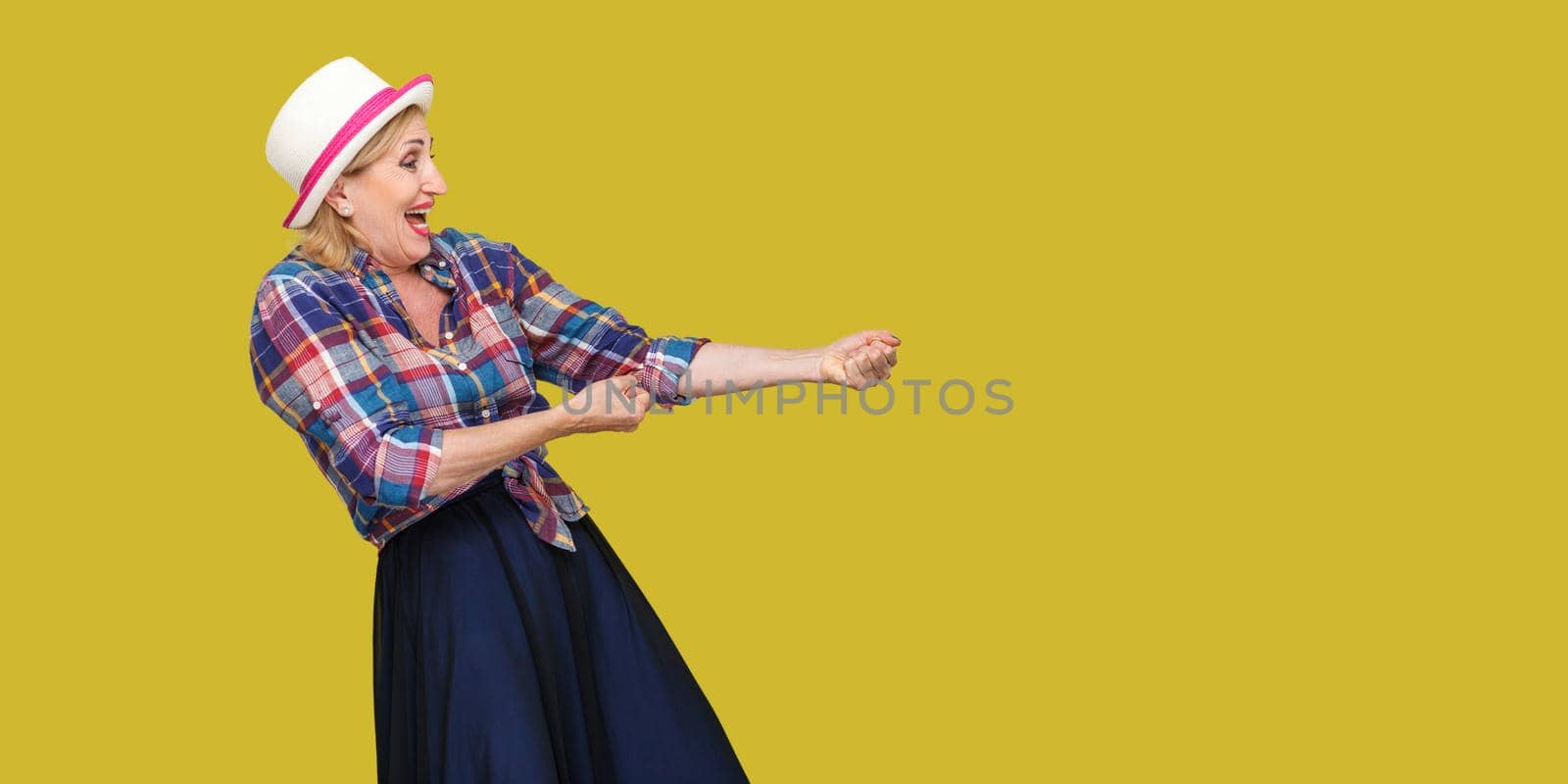 Profile side view portrait of seroius stylish mature woman in casual style with hat standing in attack mode or doing pulling gesture of something. indoor studio shot isolated on yellow background.