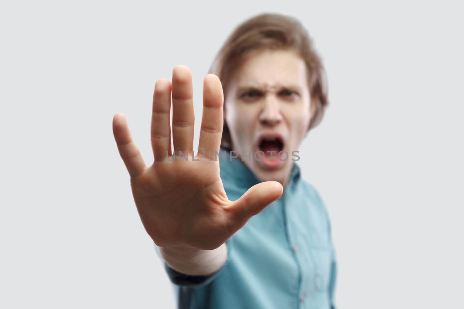 Stop it. Portrait of angry handsome long haired blonde man in blue shirt standing with stop hand gesture sign and looking at camera screaming. indoor studio shot, isolated on light grey background.