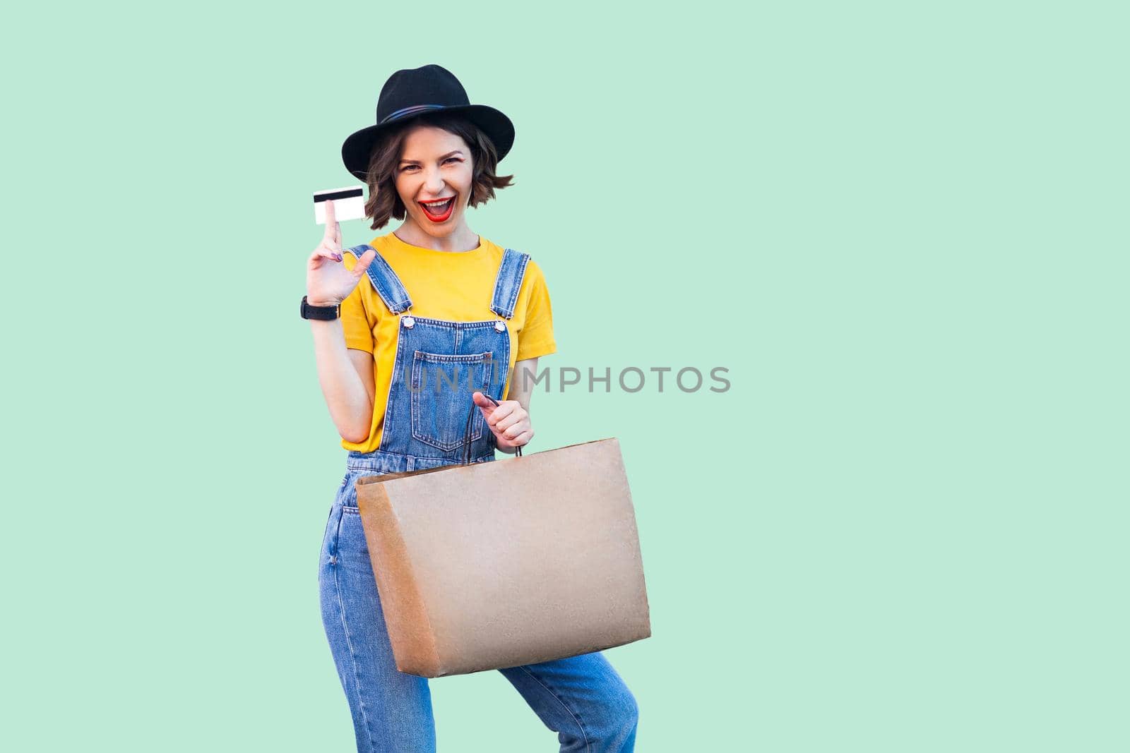 Pleasure beautiful young girl in hipster wear in denim overalls and black hat standing, holding bank card and shopping bag, open mouth, shopping online, looking at camera. Studio shot,green background