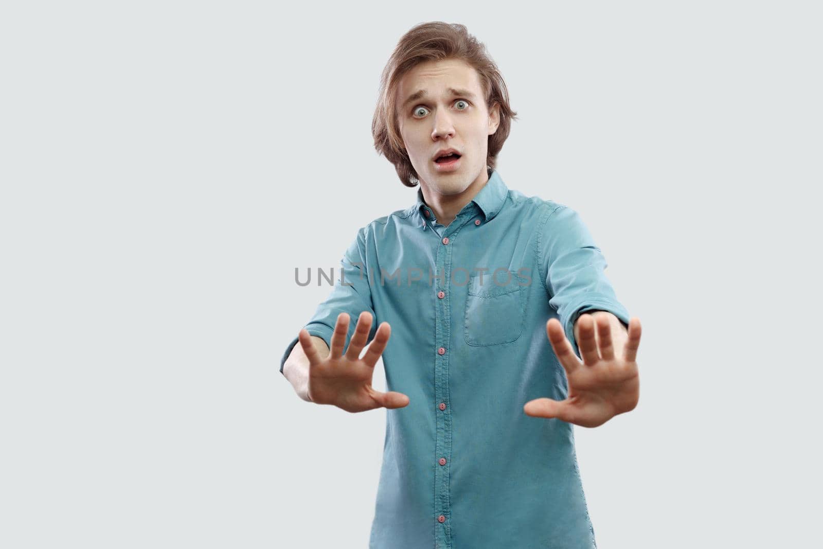 Portrait of scared or shocked handsome long haired blonde young man in blue casual shirt standing with blocking hand and looking at camera. indoor studio shot, isolated on light grey background.