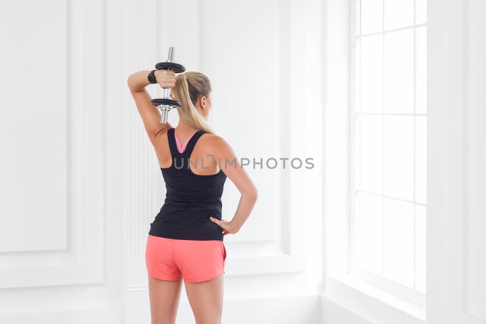 Striving for strong. Backside of young adult motivation athletic bodybuilder woman in pink shorts and black top standing, holding dumbells doing exercise for arms at the gym with perfect figure.