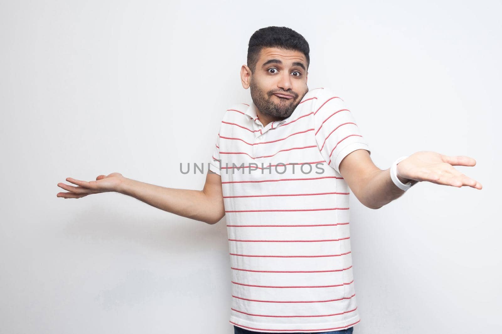 I don't know. Portrait of confused handsome bearded young man in striped t-shirt standing with raised arms and dont know what to do. indoor studio shot, isolated on white background.