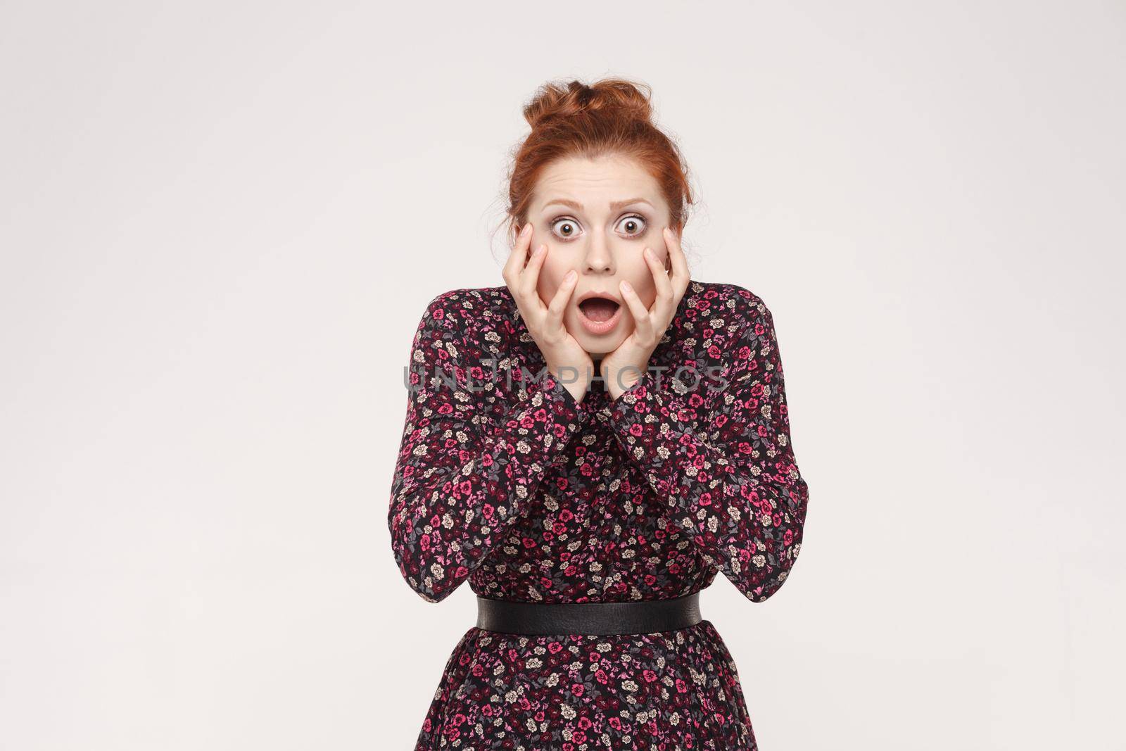 Human face expressions and emotions. Redhead woman screaming with shock, holding hands on her cheeks. Isolated studio shot on gray background.