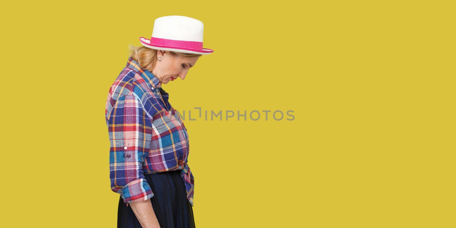 Profile side view portrait of sad alone stylish mature woman in casual style with hat standing, holding head down, frowning and crying. indoor studio shot isolated on yellow background.