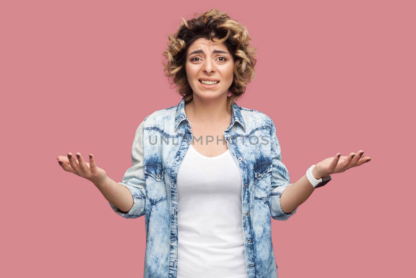 Portrait of confused or shocked young woman with curly hairstyle in casual blue shirt standing with raised arms and looking at camera worried. indoor studio shot, isolated on pink background.