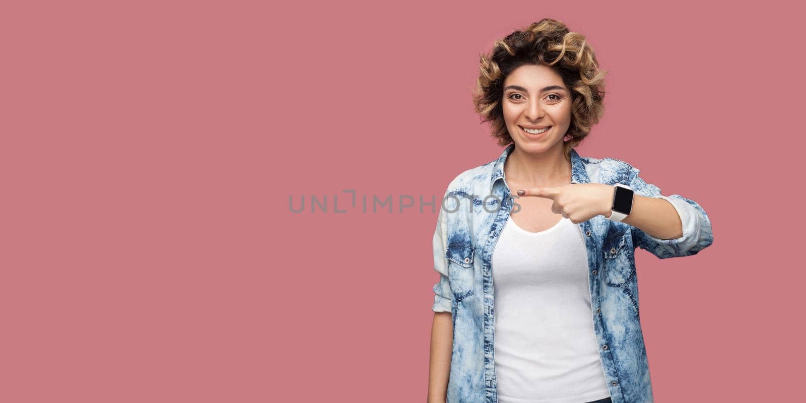 Portrait of young happy woman with curly hairstyle in casual blue shirt standing, toothy smile and pointing at background empty copyspace. indoor studio shot, isolated on pink background.