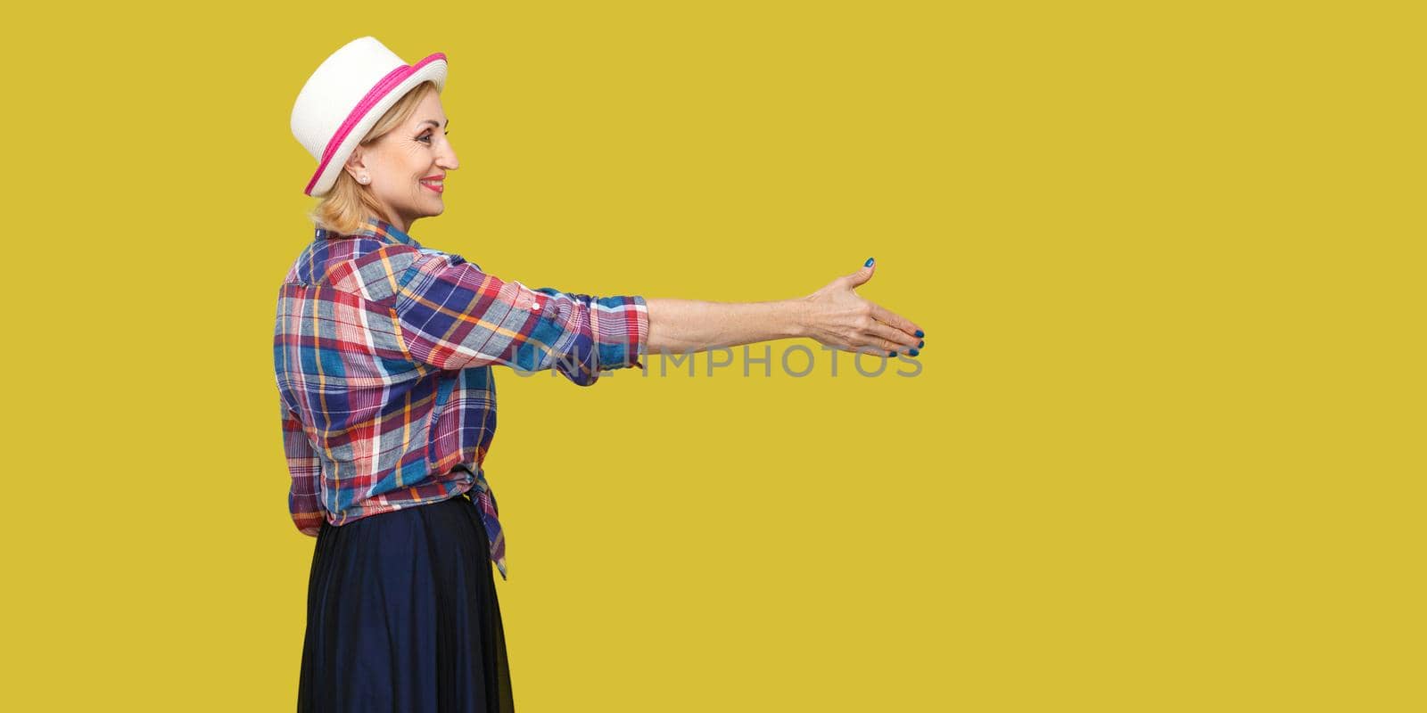 Profile side view portrait of happy modern stylish mature woman in casual style with hat standing, looking with toothy smile, giving hand to greeting. indoor studio shot isolated on yellow background.