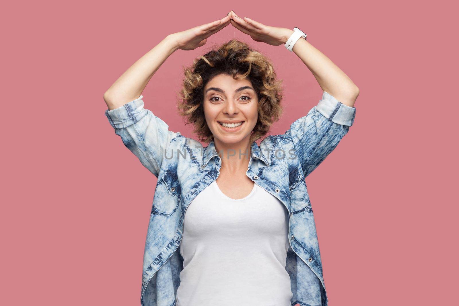 Portrait of happy young woman with curly hairstyle in casual blue shirt standing with roof gesture hands on head and looking at camera with toothy smile. indoor studio shot isolated on pink background
