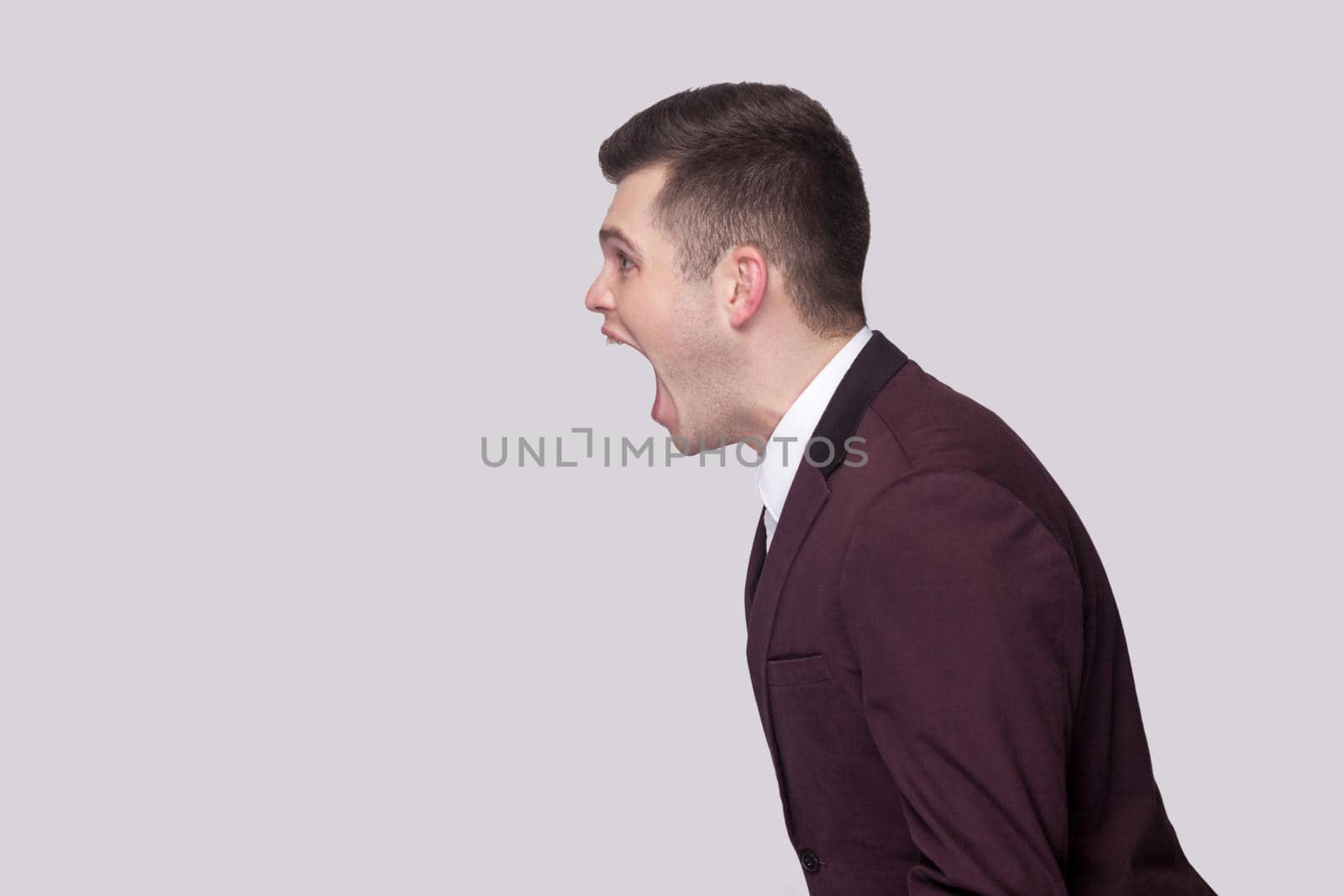 Profile side view portrait aggressive of handsome young man in violet suit and white shirt, standing and screaming with nervous face. indoor studio shot, isolated on grey background.