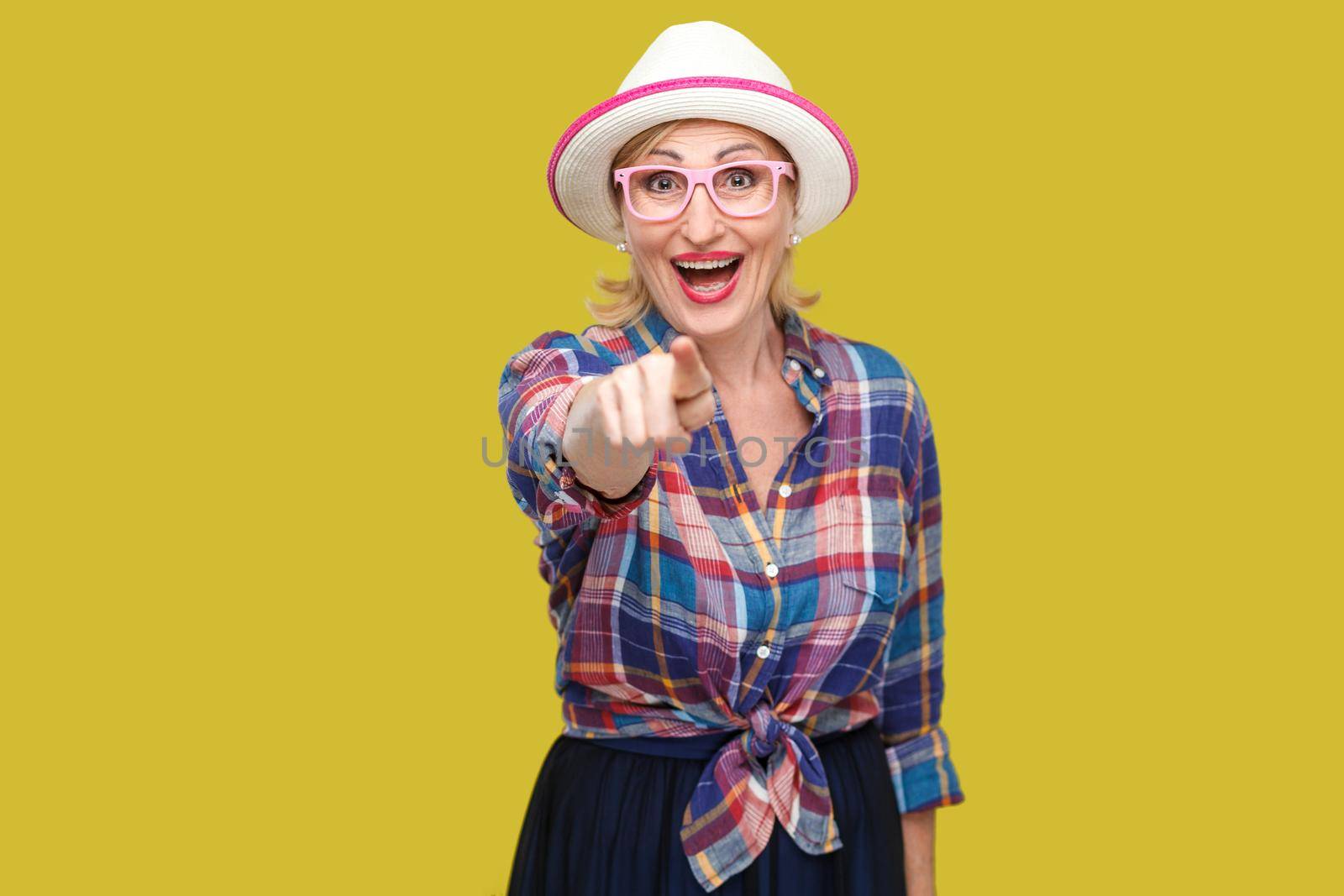 Portrait of funny surprised modern stylish mature woman in casual style with hat and eyeglasses standing, looking and pointing at camera with amazed face. studio shot isolated on yellow background.