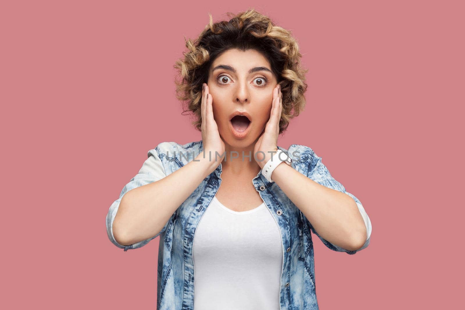 Portrait of shocked young woman with curly hairstyle in casual blue shirt standing with big eyes and open mouth and looking at camera surprised. indoor studio shot, isolated on pink background.