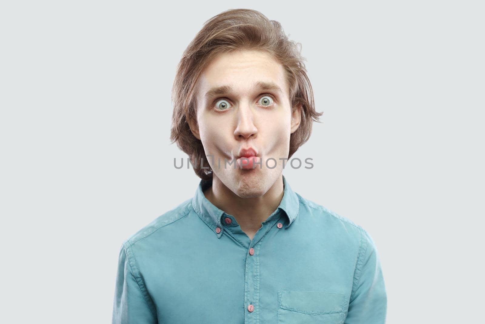 Portrait of funny handsome long haired blonde young man in blue casual shirt standing, looking at camera with big eyes and making fish lips. indoor studio shot, isolated on light grey background.