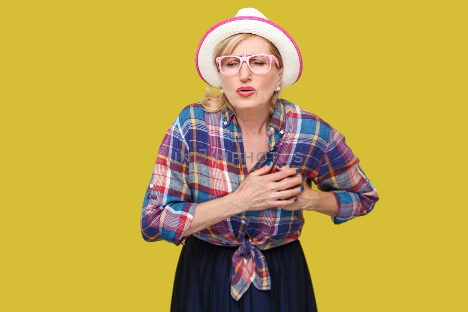 Heart attack pain or infarct. Portrait of mature woman in casual style with hat, eyeglasses standing, holding her breast because feeling pain on heart. indoor studio shot isolated on yellow background