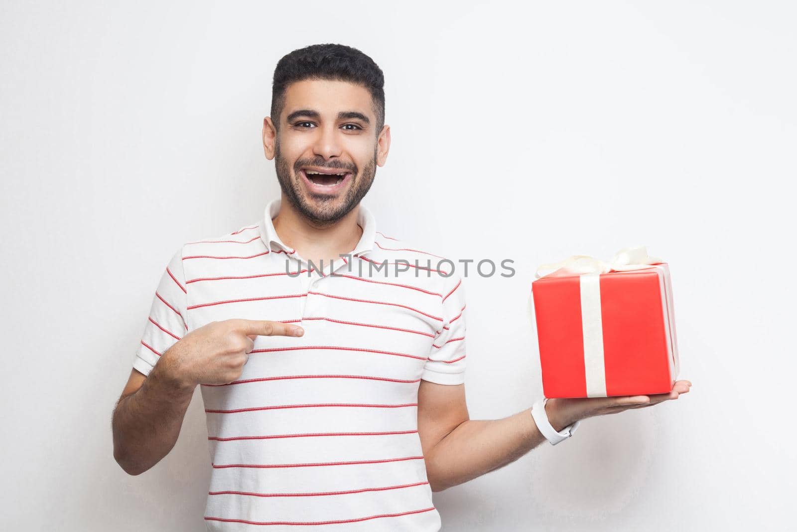 It is yours? Satisfied handsome modern young adult man in t-shirt standing, holding big gift box with opened mouth, pointing finger, looking at camera. Studio shot, white background, isolated, indoor