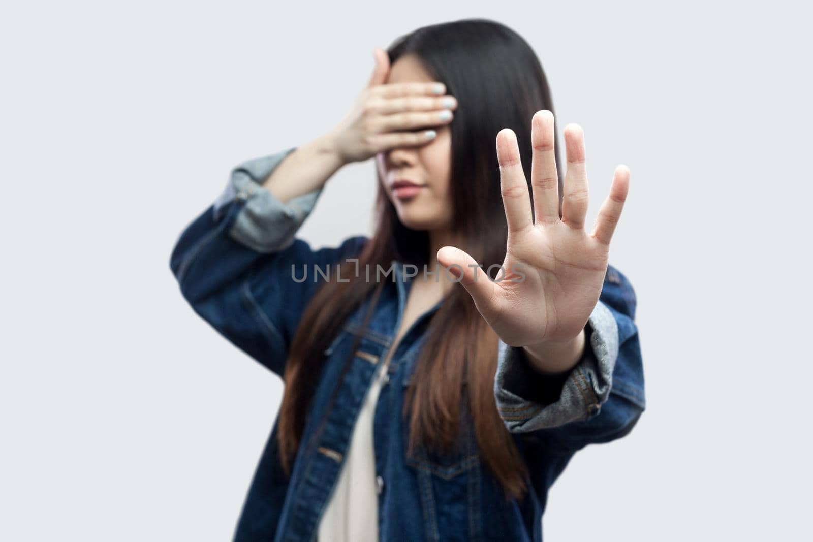Stop, I dob't want to see this. Portrait of scared brunette young woman in casual blue denim jacket standing closed eyes and showing stop gesture. indoor studio shot, isolated on light grey background