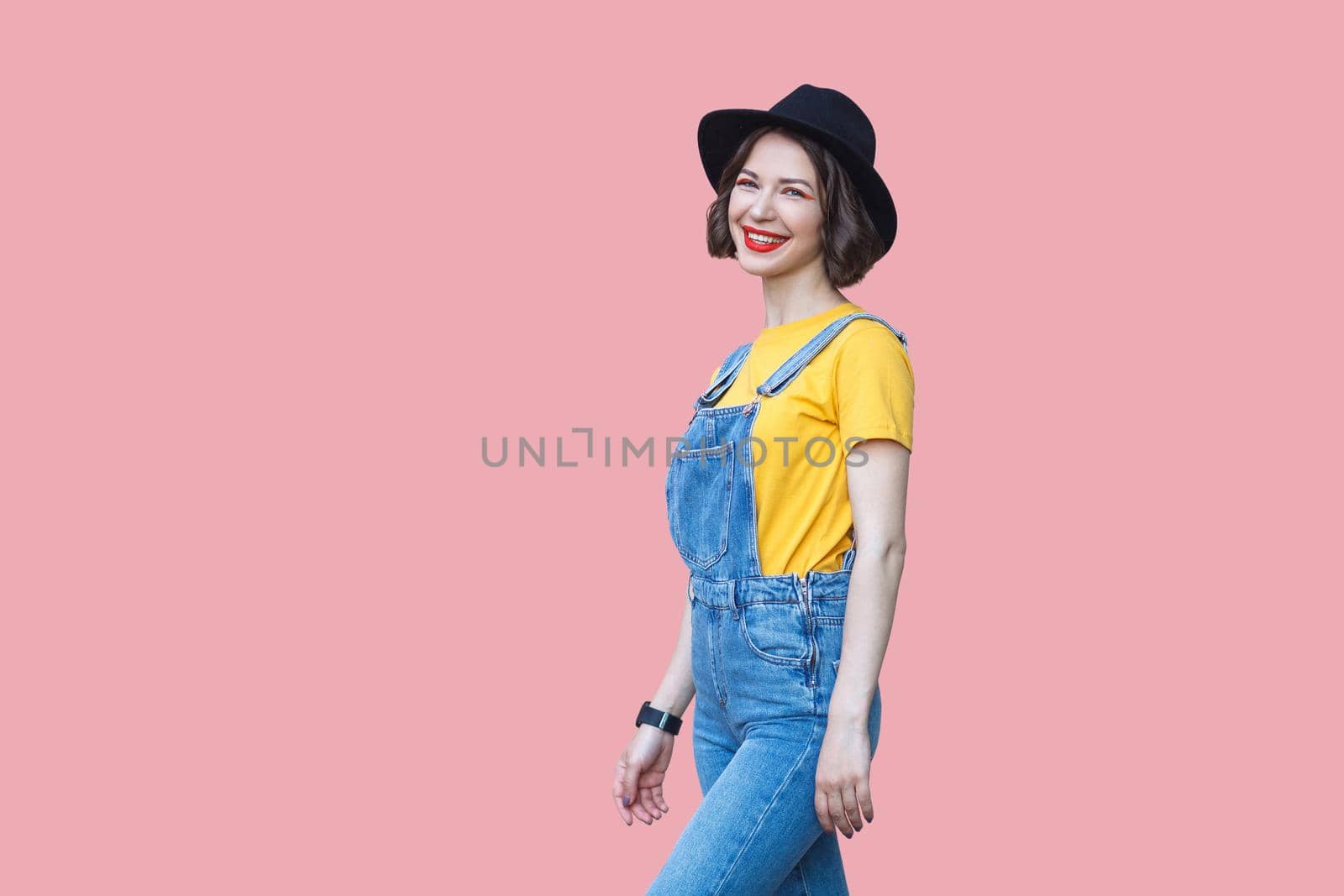 Portrait of happy beautiful young woman in yellow t-shirt and blue denim overalls with makeup and black hat standing and looking at camera with toothy smile. studio shot isolated on pink background.