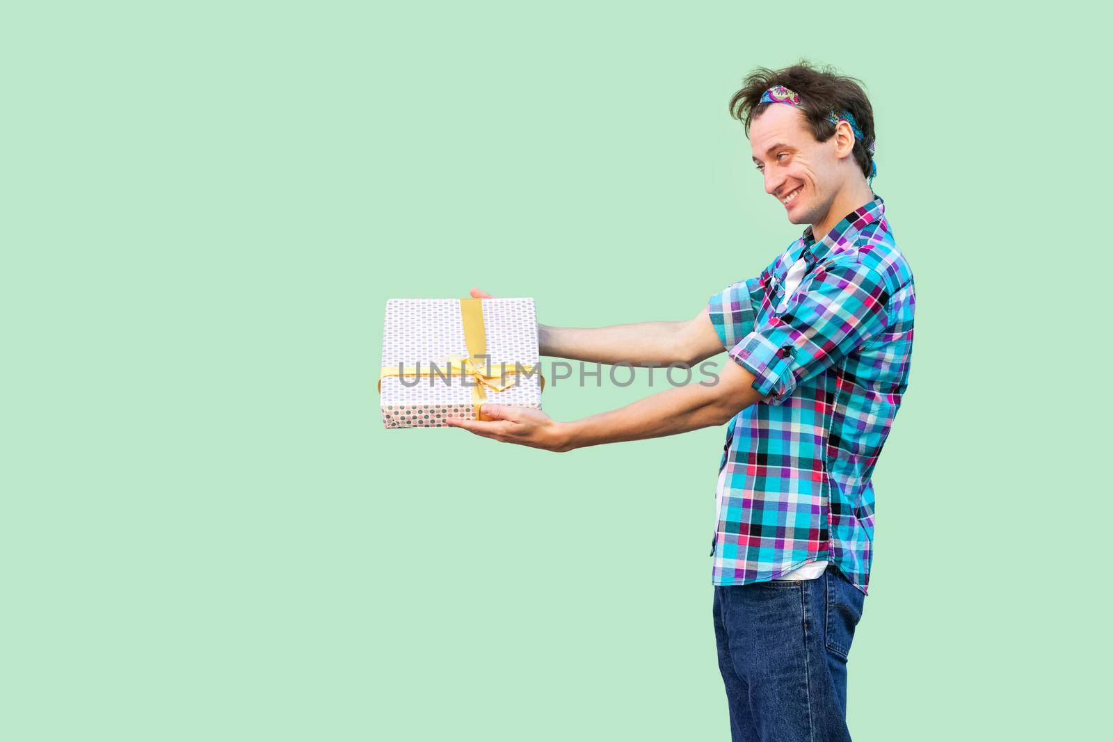 Side view of happy young hipster man in white t-shirt and checkered shirt standing, giving you present with yellow bow, toothy smile. Indoor, copy space, isolated, studio shot, green background