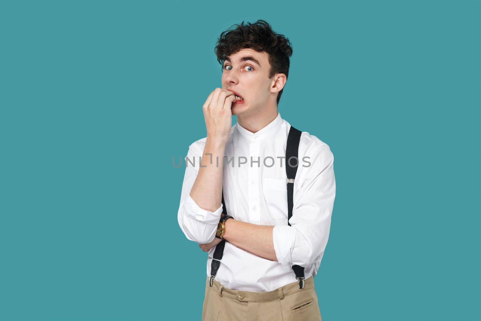 Nervous man bitting nails and looking at camera. Portrait of handsome hipster curly young businessman in classic casual white shirt, suspender standing. indoor studio shot isolated on blue background.