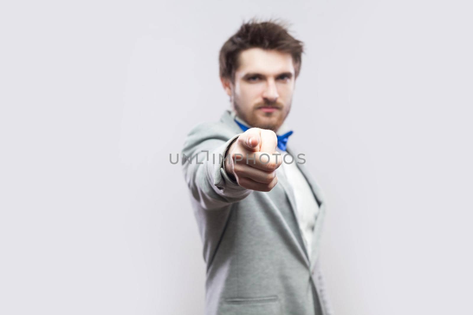Portrait of serious handsome bearded man in casual grey suit and blue bow tie standing and looking and blaming at camera. indoor studio shot, isolated on light grey background.