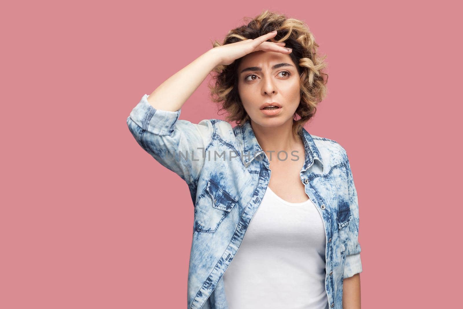 Portrait of attentive young woman with curly hairstyle in casual blue shirt standing with hand on forehead and looking away with serious face and attention. studio shot, isolated on pink background.