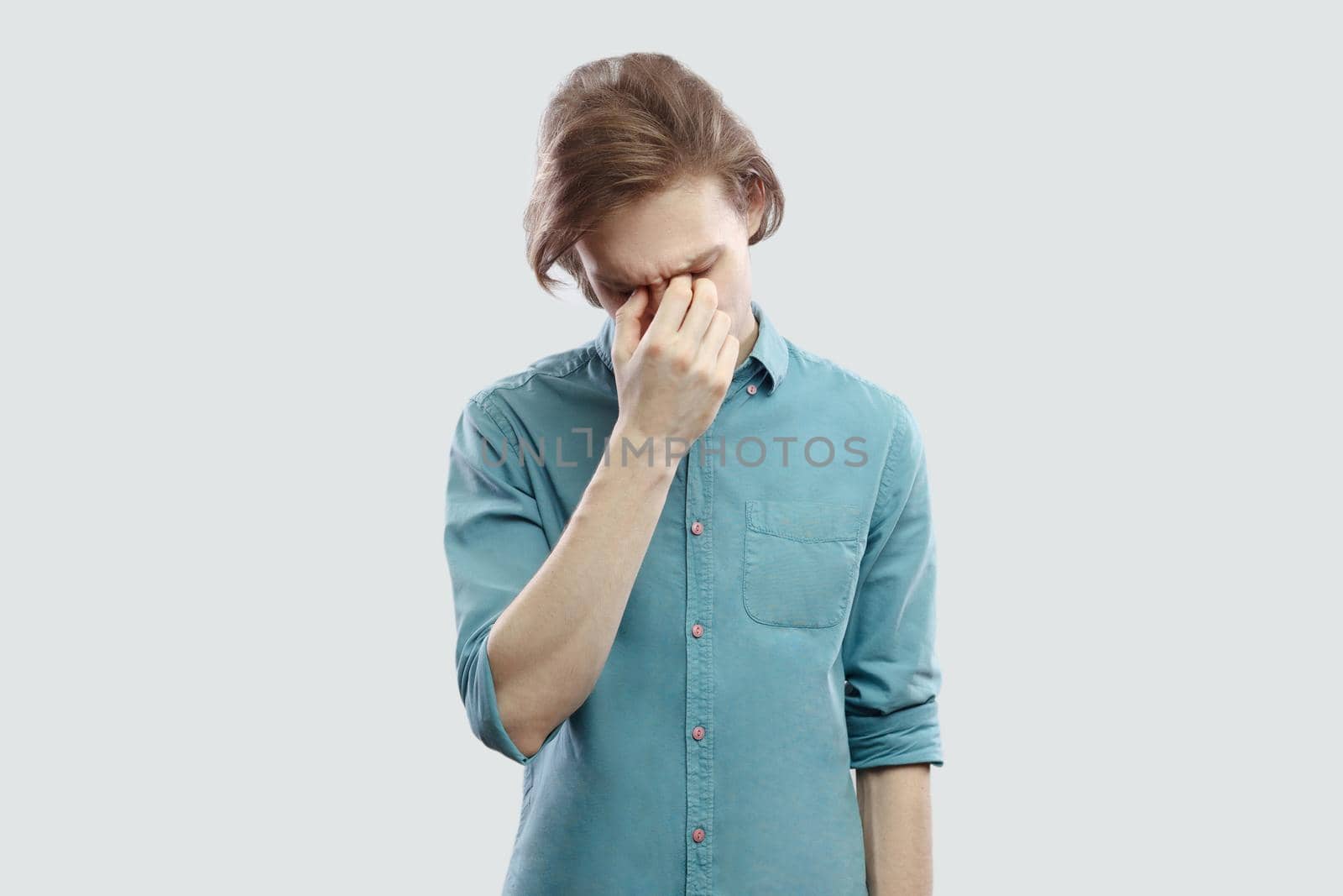 Portrait of sad alone handsome long haired blonde young man in blue casual shirt standing, holding his head down and crying. indoor studio shot, isolated on light grey background.