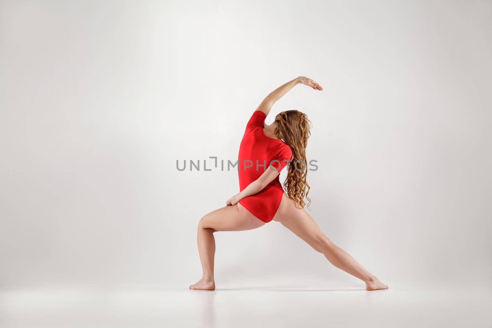 athletic blonde woman in red leotard doing yoga urdhva virabhadrasana pose . isolated indoor studio shot on light gray background. healthy lifestyle and leisure activity concept.