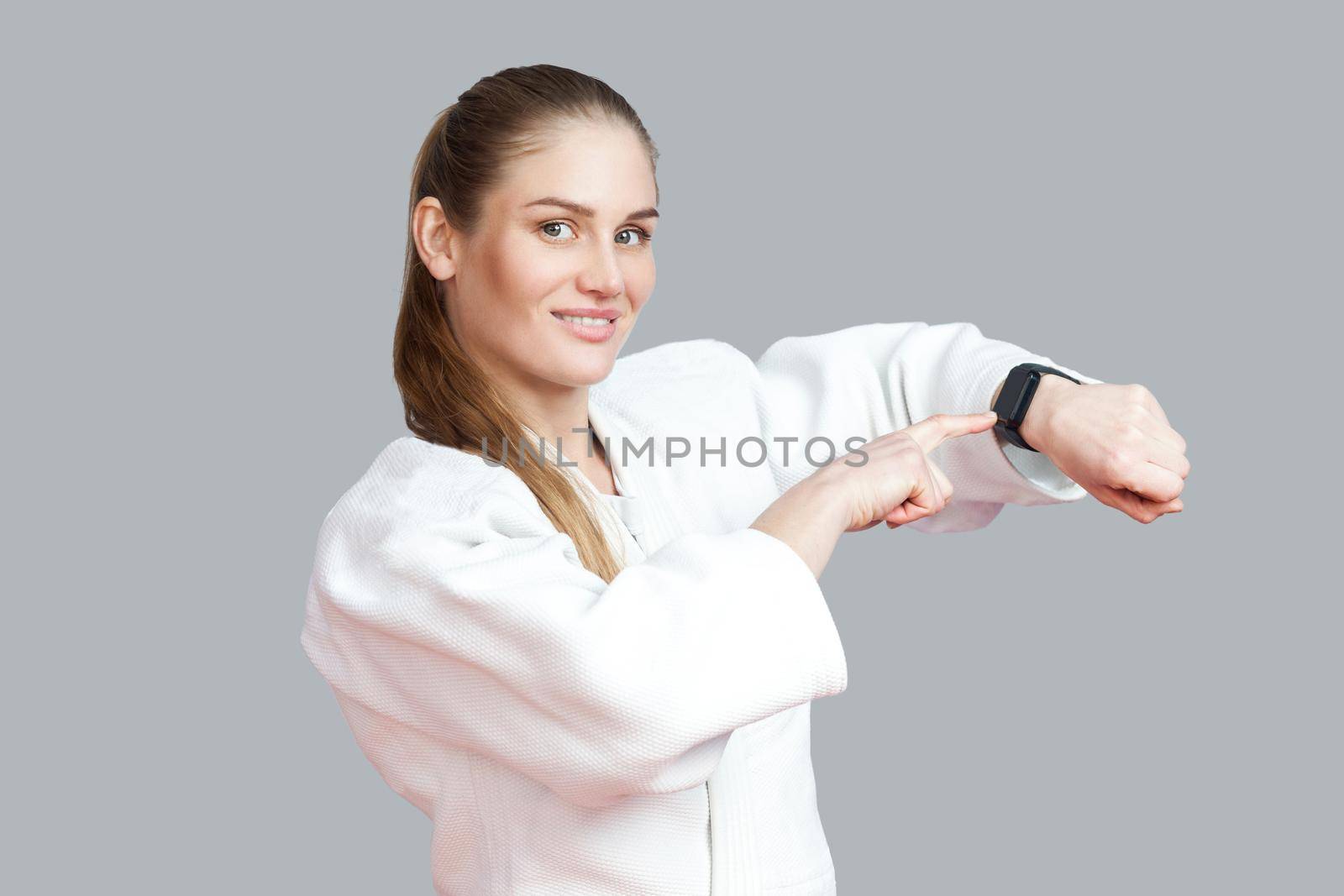 Burning calories. Beautiful young blond athletic woman pointing finger at smart watch, toothy smile and looking at camera. Woman using fitness bracelet. Indoor studio shot, isolated on grey background
