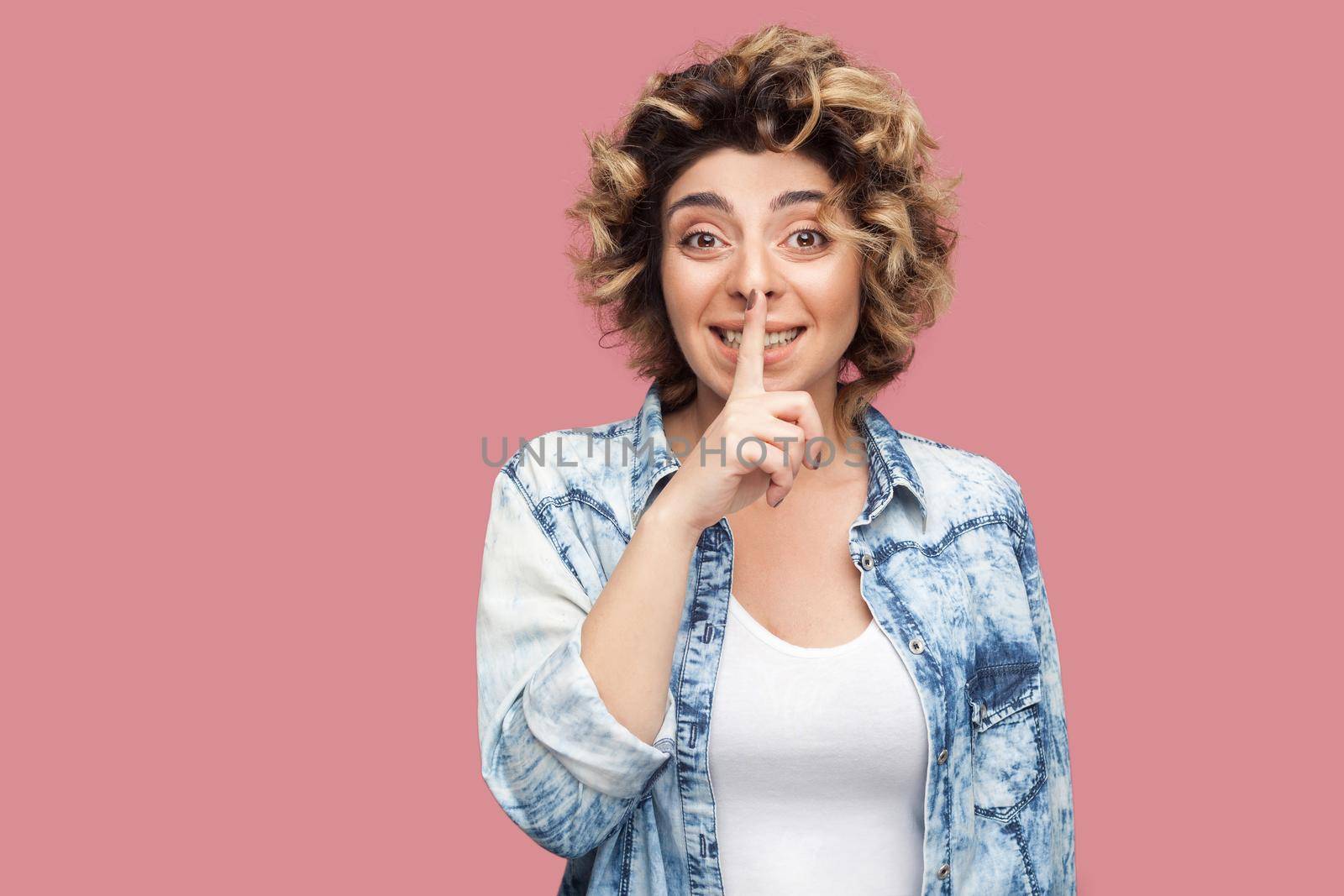 Portrait of worry young woman with curly hairstyle in casual blue shirt standing, telling secret and looking at camera with finger on lips secret sign. indoor studio shot, isolated on pink background.