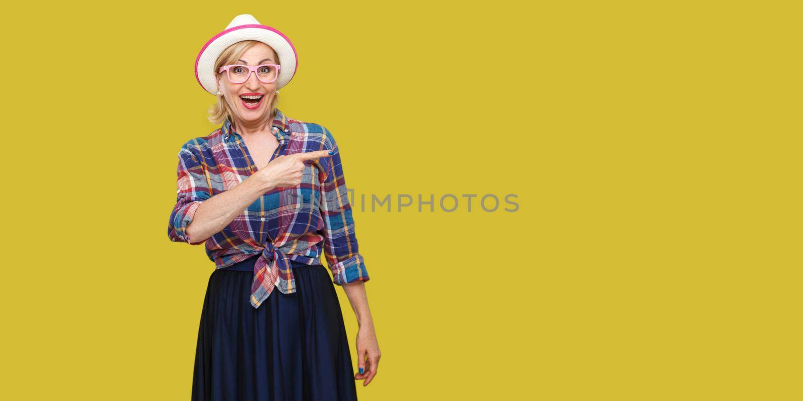 Portrait of surprised modern stylish mature woman in casual style with hat and eyeglasses standing, amazed looking at camera and pointing at wall. indoor studio shot isolated on yellow background.