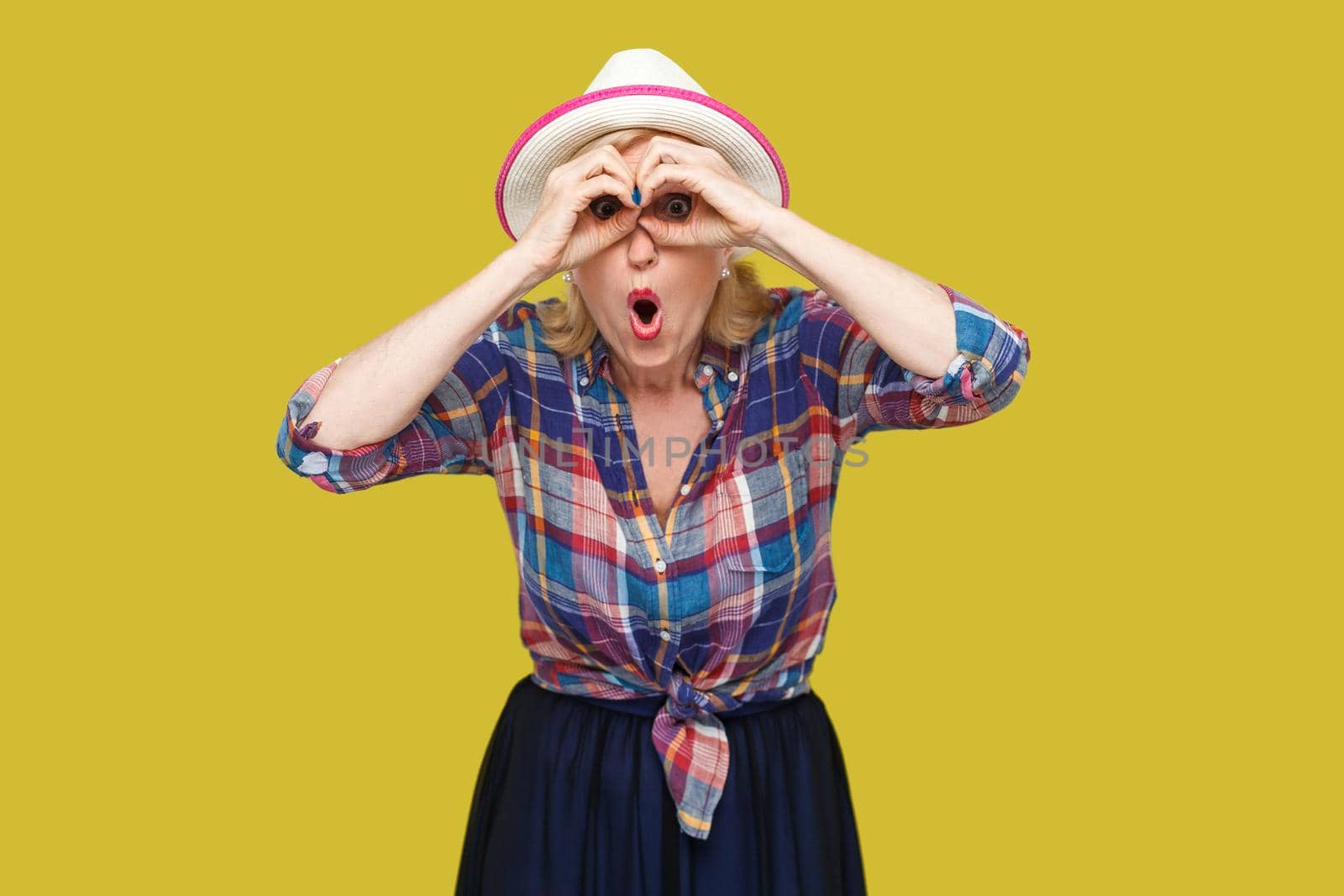 Portrait of funny surprised modern stylish mature woman in casual style with hat standing with binoculars gesture and looking with amazed face. indoor studio shot isolated on yellow background.