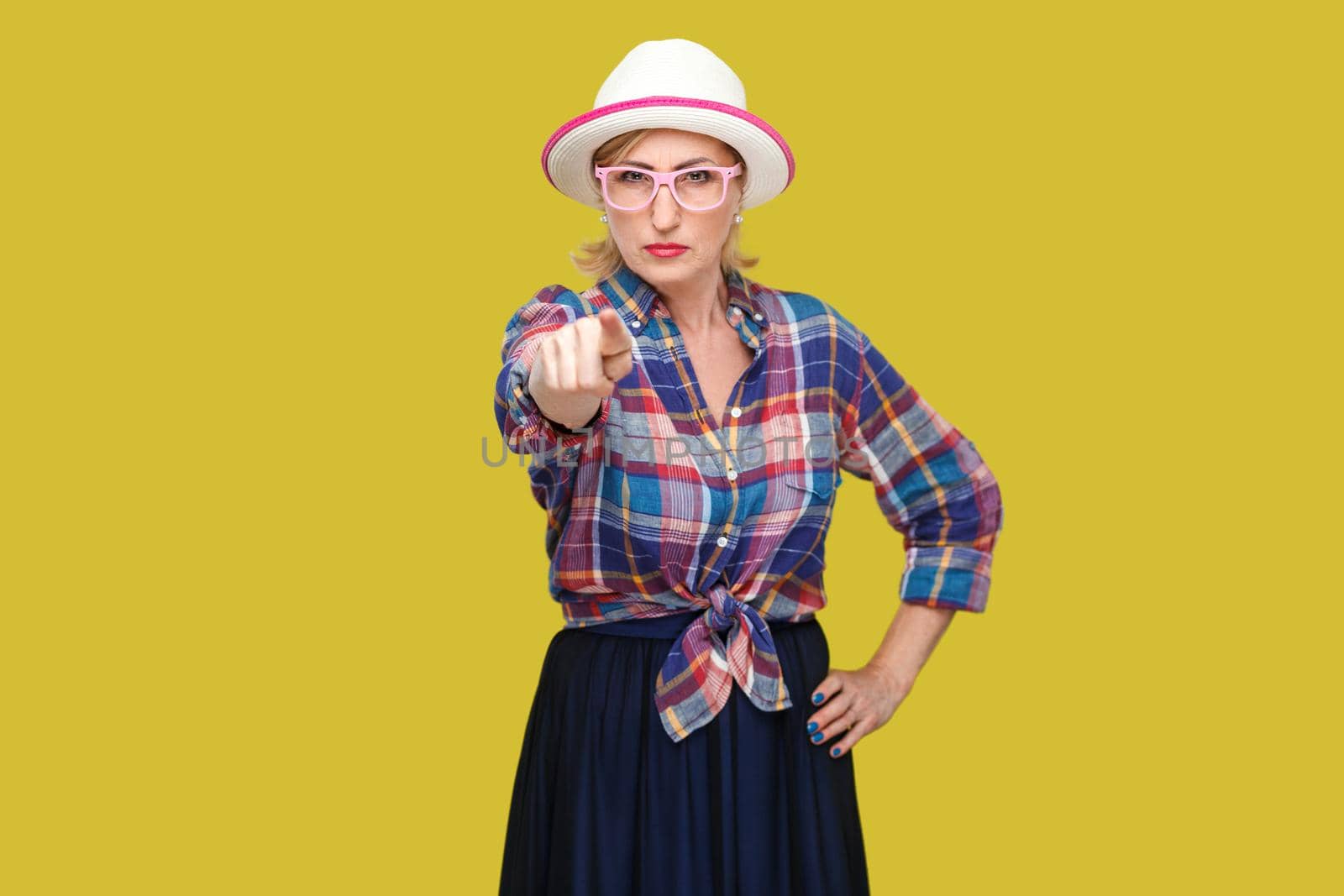 Hey you. Portrait of serious bossy modern stylish mature woman in casual style with hat and eyeglasses standing, pointing and looking at camera. indoor studio shot isolated on yellow background.