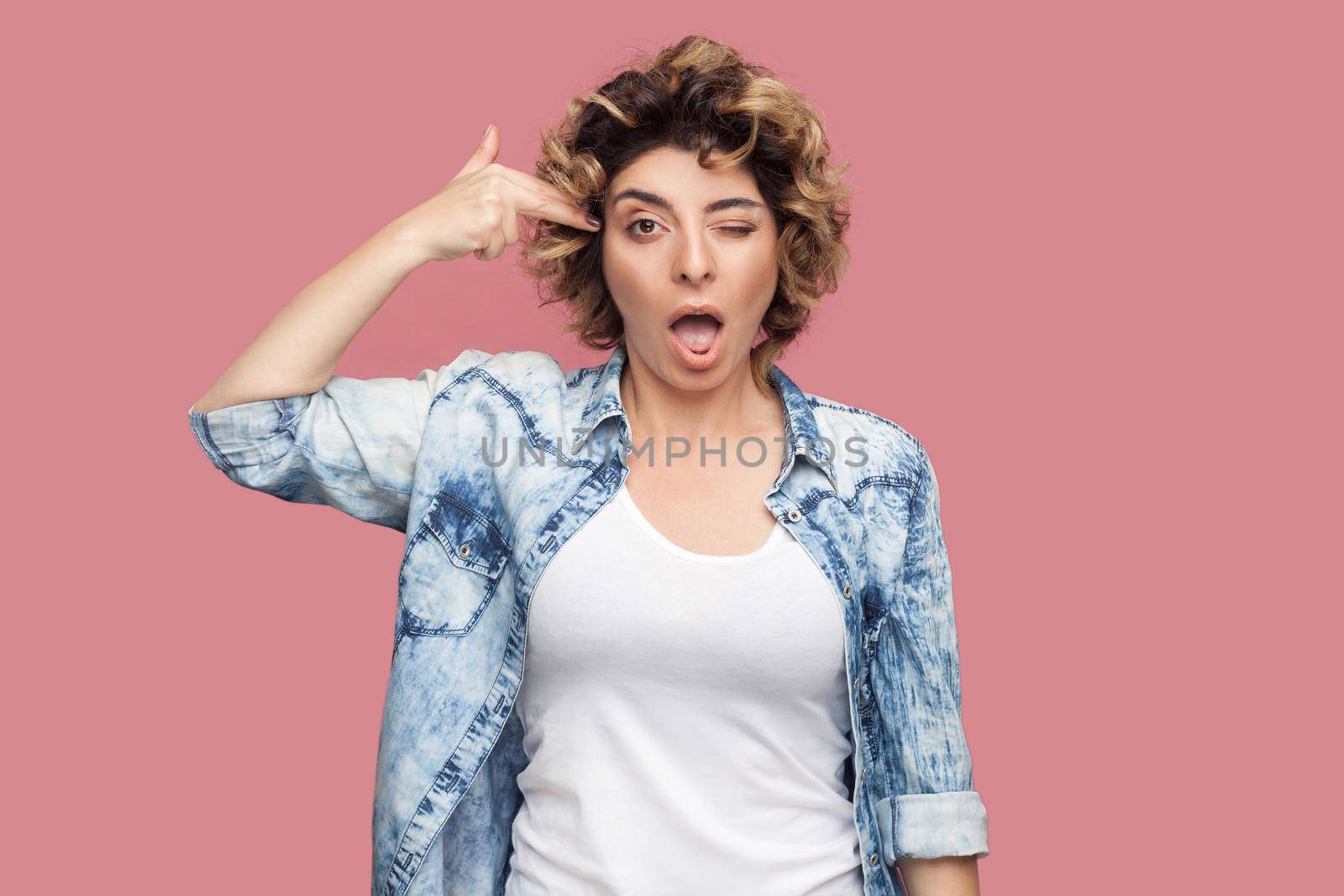 Portrait of crazy young woman with curly hairstyle in casual blue shirt standing with pistol hand gun on her head and looking at camera. indoor studio shot, isolated on pink background.
