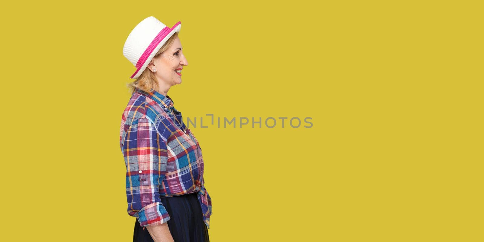 Profile side view portrait of happy successful modern stylish mature woman in casual style with hat standing, looking forward with toothy smile. indoor studio shot isolated on yellow background.