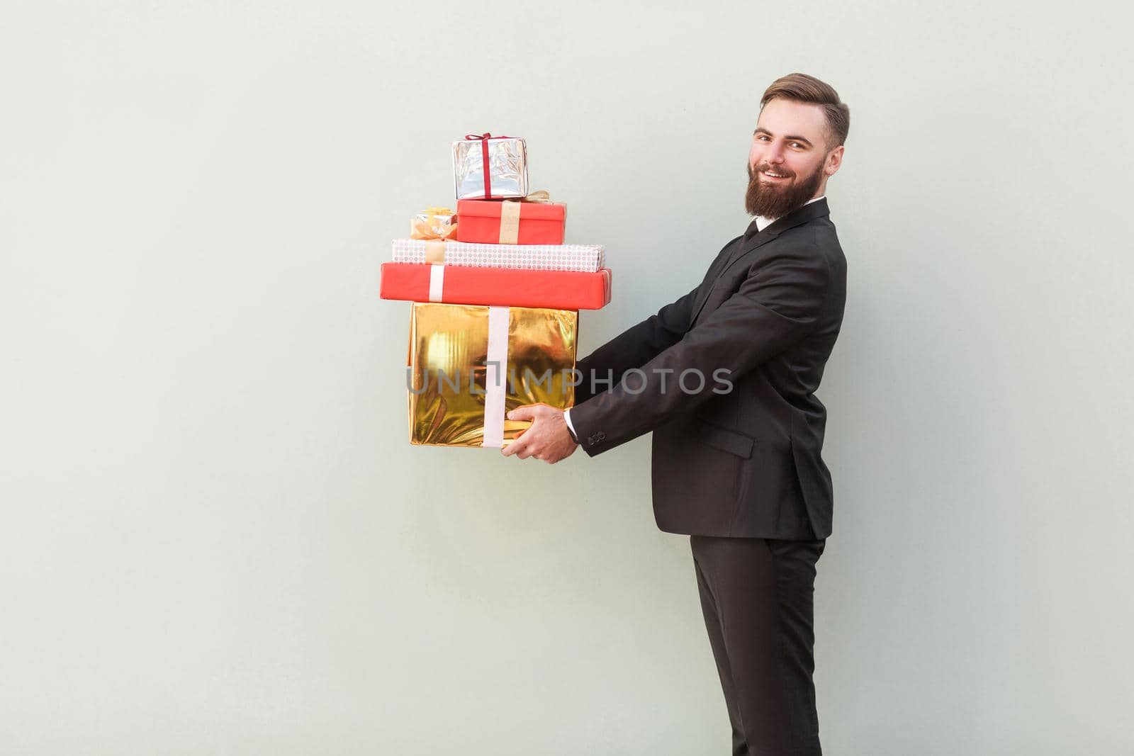 Profile, side view businessman toothy smile and holding gift boxes. Indoor shot, gray background