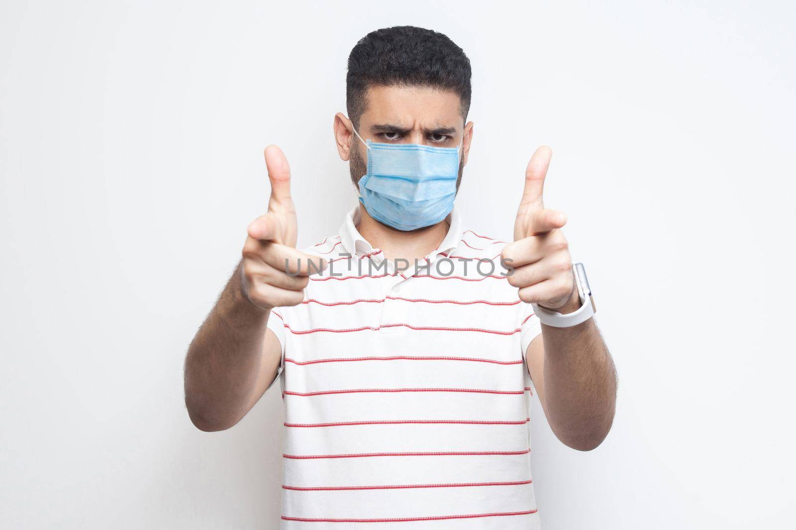 Hey you. Portrait of serious bossy young man with surgical medical mask in striped t-shirt standing, looking and pointing at camera. indoor studio shot, isolated on white background.