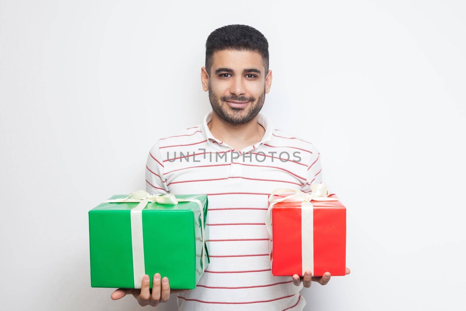 Portrait of positive attractive young adult man in t-shirt standing, holding two big gift box, looking at camera. Studio shot, white background, isolated, indoor, copy space