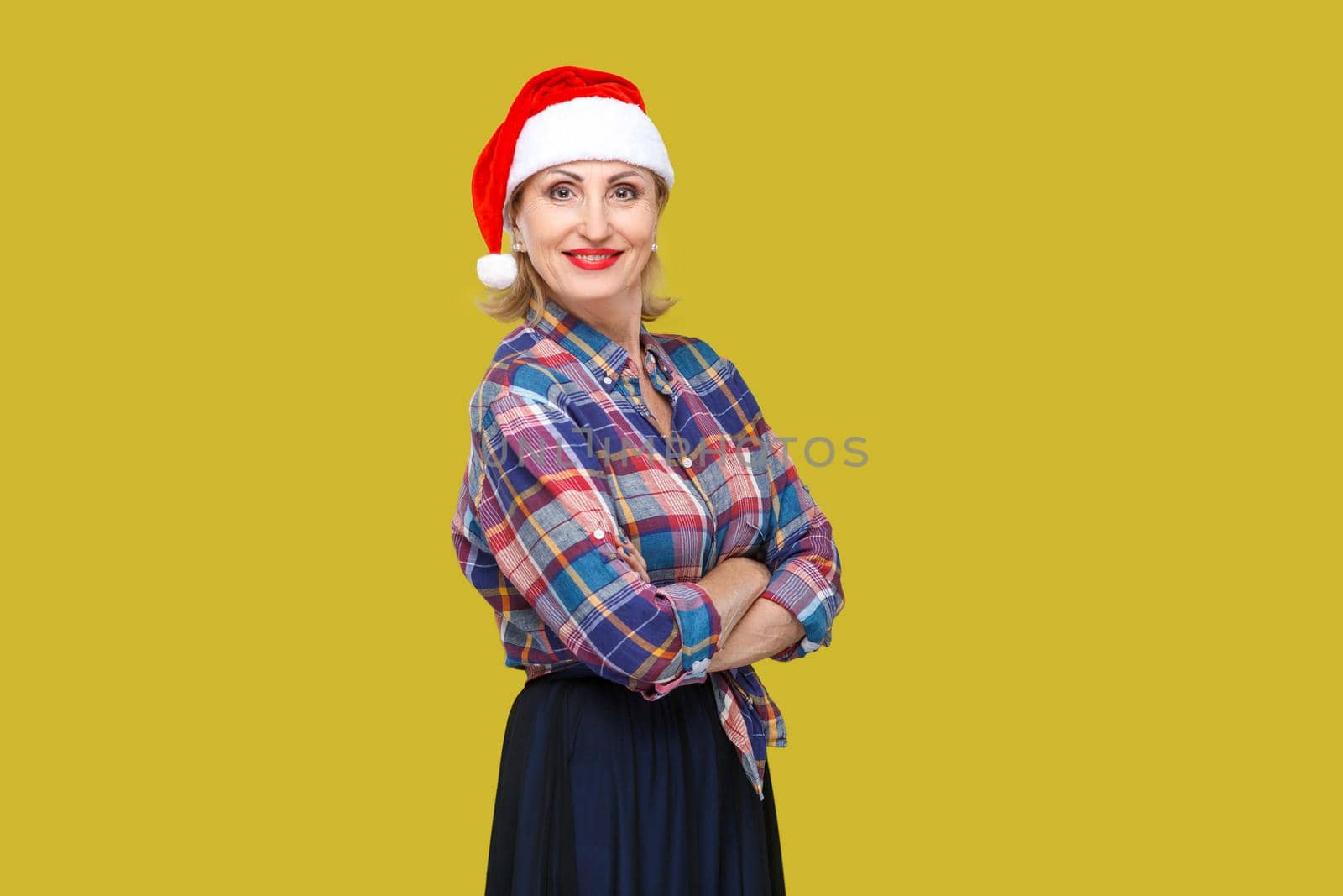 Portrait of confident attractive adult woman in red santa cap and checkered shirt standing and closed up hands with toothy smiling, looking at camera. Indoor, studio shot, yellow background