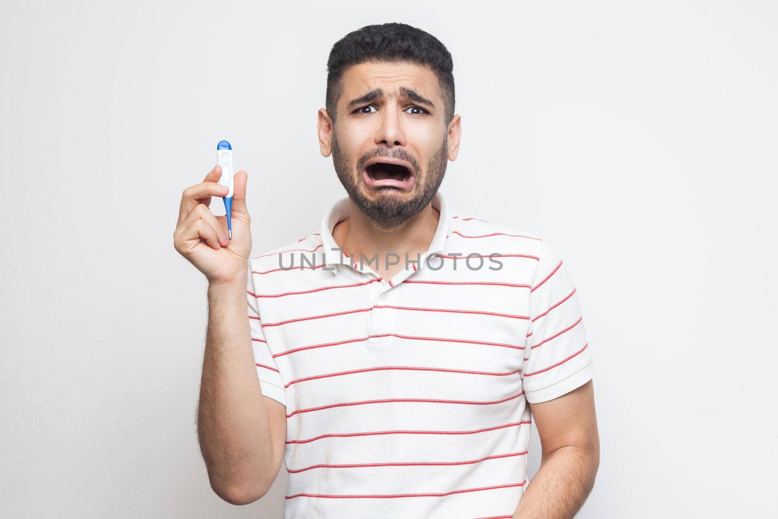 Scared worry sick young adult man in white t-shirt standing and holding thermometer are shoched for high temperature, looking at camera. Indoor, isolated, white background, studio shot